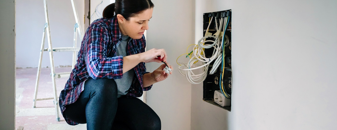 A woman attempts to fix exposed wires in a wall, posing a potential electrical hazard during home renovation.