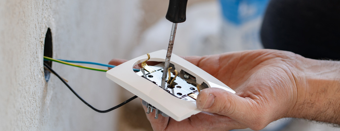 A residential electrician installs a wall socket, connecting colored wires using a screwdriver.