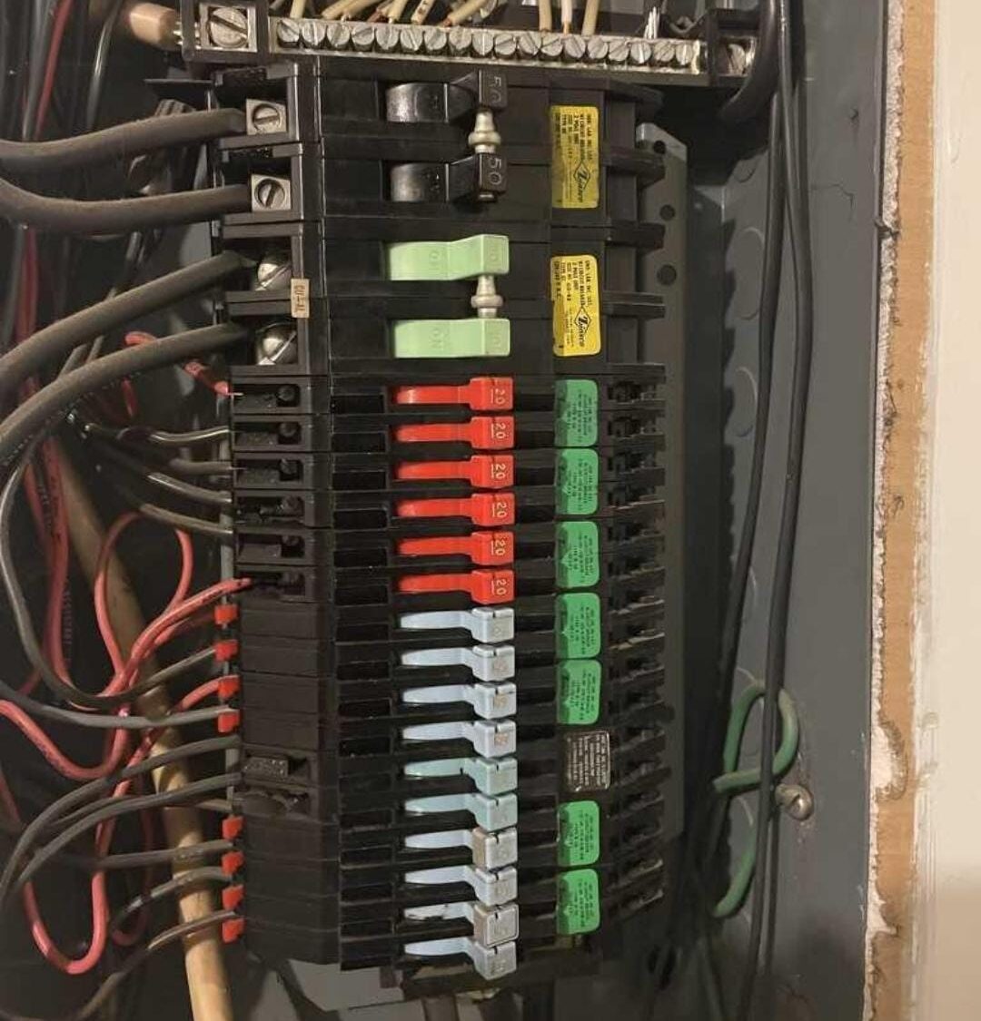 Interior of a residential electrical panel with numerous circuit breakers and tangled wires at the top and sides.